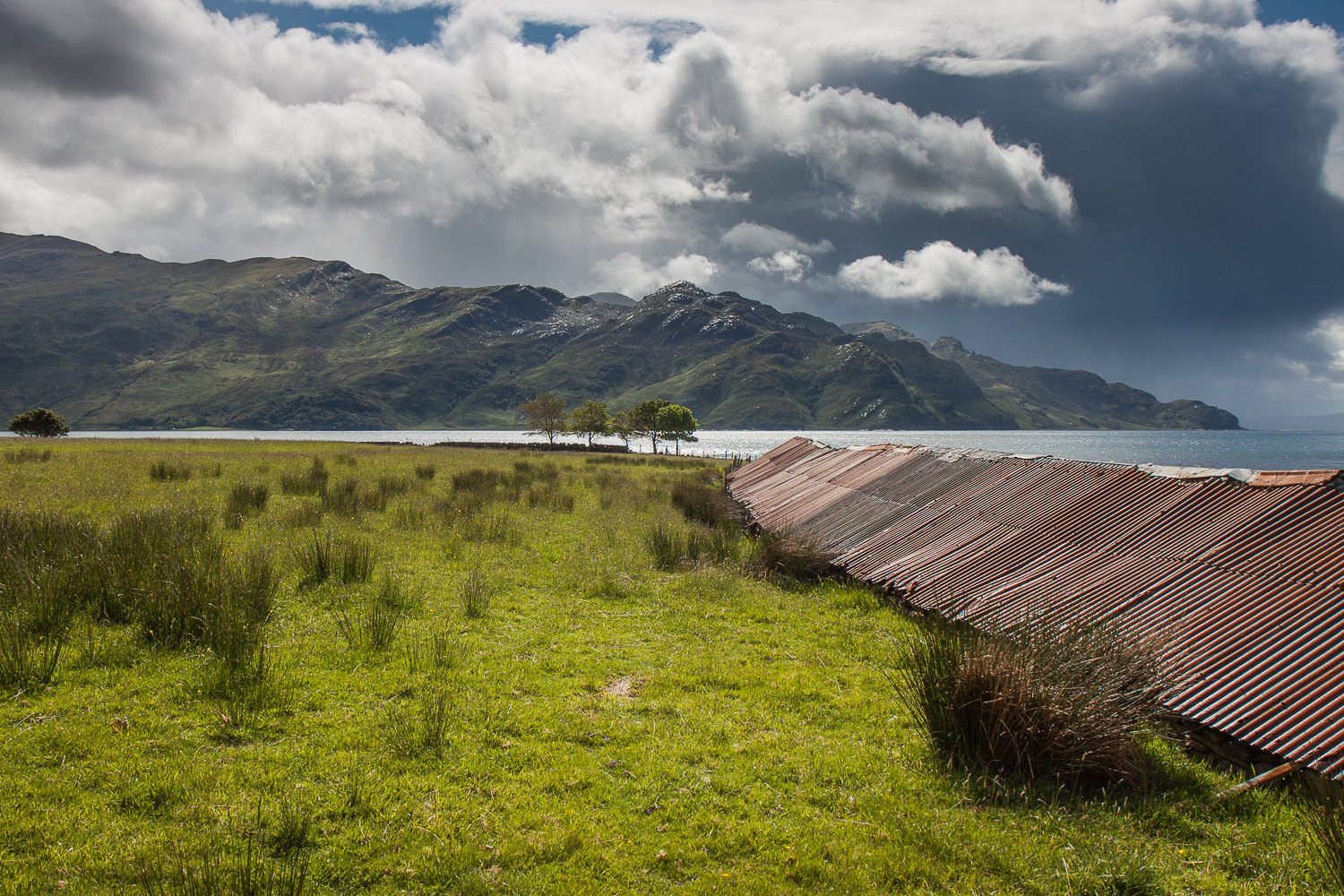 Lange schuur met roestende golfplaten als dak, heuvels en wolken in de achtergrond.